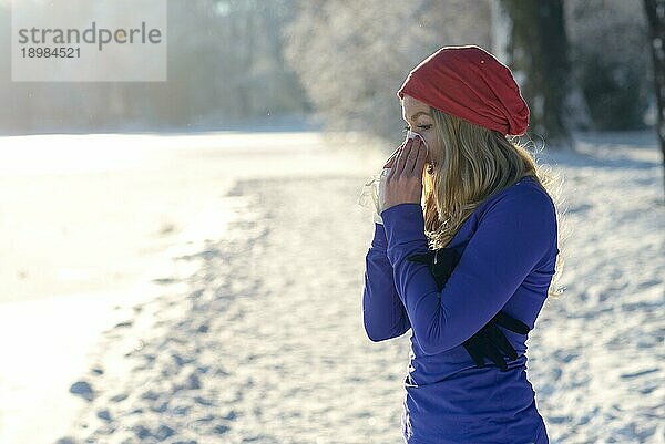 Junge Frau  die an einem Winterschnupfen leidet  steht im Freien in einer verschneiten kalten Landschaft und schnäuzt sich die Nase mit einem Taschentuch  mit Kopierraum über unberührtem weißen Schnee