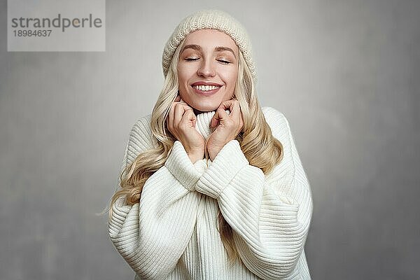 Junge blonde Frau  die sich an ihrem weißen Strickpulli erfreut  steht vor einem grauen Hintergrund  hält die Hände in den Nacken und lächelt mit geschlossenen Augen