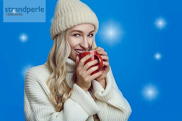 Junge Frau  die eine Tasse heißen Kaffee im Winter genießt und ihn in ihren Händen schröpft  während sie mit einem glückseligen Lächeln vor einem bläulichen Hintergrund trinkt