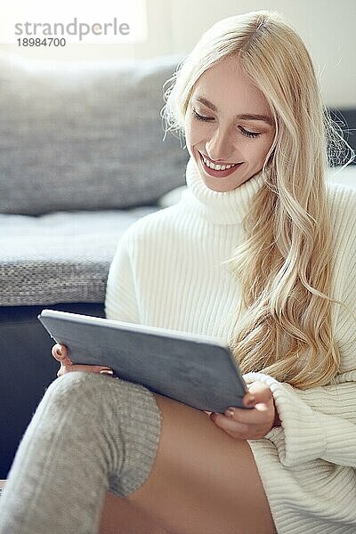 Trendy lächelnde blonde Frau in Winterleggings und einem warmen Wollpullover  die sich zu Hause mit einem Tablet PC entspannt und mit einem schönen  lebhaften Lächeln liest