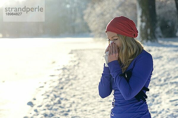 Junge Frau  die an einem Winterschnupfen leidet  steht im Freien in einer verschneiten kalten Landschaft und schnäuzt sich die Nase mit einem Taschentuch  mit Kopierraum über unberührtem weißen Schnee