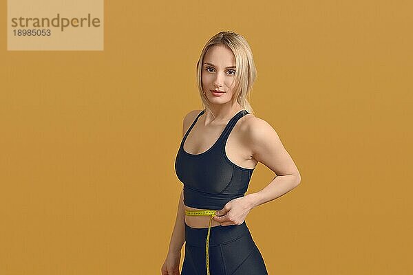Schlanke attraktive junge blonde Frau  die ihre Taille mit einem Maßband mißt  um sich gesund zu ernähren  Sport zu treiben und ein Fitnesskonzept zu verfolgen  auf einem gelben Hintergrund mit Leerzeichen