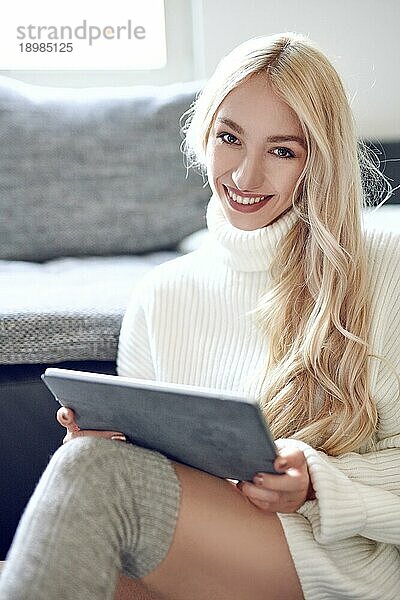 Trendy lächelnde blonde Frau in Winterleggings und einem warmen Wollpullover  die sich zu Hause mit einem Tablet PC entspannt und mit einem schönen  lebhaften Lächeln in die Kamera schaut