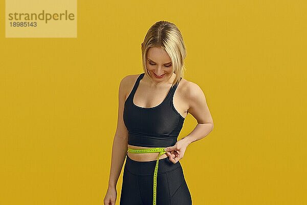 Schlanke  sportliche junge Frau  die ihre Taille mit einem Maßband mißt  während sie mit einem zufriedenen Lächeln nach unten blickt  in einem Konzept für Gewichtsabnahme  gesunde Ernährung und Bewegung auf einem gelben Hintergrund mit Kopierraum
