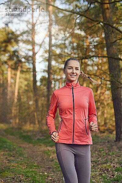 Gesunde  fitte  attraktive junge Frau  die im Wald auf einem Waldweg joggt und sich der Kamera mit einem glücklichen Lächeln nähert  in einem aktiven Lebensstil  Gesundheits und Fitnesskonzept