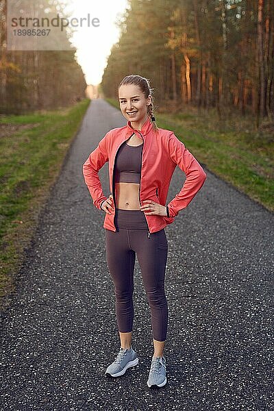 Frontalporträt in voller Länge eines sportlichen jungen Mädchens in Trainingskleidung mit grauer Hose und roter Jacke  das nach dem Joggen steht und sich ausruht  die Hände in die Hüften gestemmt  in die Kamera schaut und lächelt