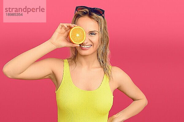 Glückliche  gesunde junge Frau  die eine halbierte  frische  saftige Orange mit einem strahlenden  freundlichen Lächeln vor einem rosafarbenen Studiohintergrund vor sich hält