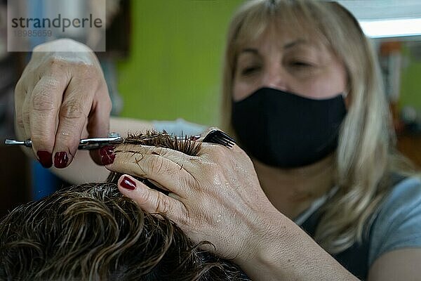 Frau mit Gesichtsmaske schneidet sich die Haare mit Schere und Kamm. Hintergrund unscharf