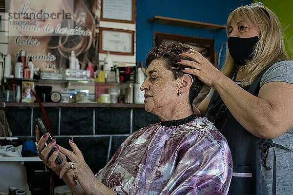 Latino Friseur Salon. Eine Kundin prüft ihr Handy und ein Friseur massiert ihr Haar. Allgemeine Aufnahme