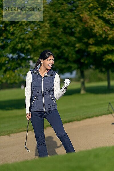 Motivierte Golferin  die eine Geste des Erfolgs und des Jubels mit der Faust macht  nachdem sie ihren Schlag ausgeführt hat  um aus einem Sandbunker auf einem Golfplatz herauszukommen