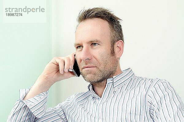 Horizontales Porträt eines gut aussehenden kaukasischen Mannes mittleren Alters  der ein langärmeliges buntes Hemd trägt  während er ein angenehmes Gespräch am Mobiltelefon führt  drinnen