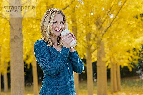 Ein schönes blondes Modell trinkt ein heißes Getränk  während es im Freien in einem Feld mit gelben Blättern posiert