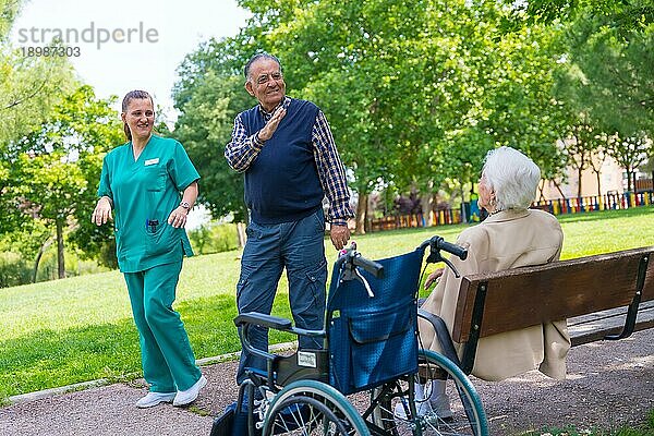Ein älterer Mann mit der Krankenschwester bei einem Spaziergang durch den Garten eines Pflegeheims  der eine ältere Frau im Rollstuhl begrüßt