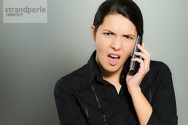 Nervöse  hübsche junge Frau  die mit ihrem Handy telefoniert und mit einem verwirrten Gesichtsausdruck und Stirnrunzeln in die Kamera schaut  vor einem grauen Studiohintergrund