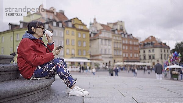 Ältere Frau sitzt auf der Treppe  trinkt Kaffee und benutzt ein Smartphone im historischen Zentrum einer alten europäischen Stadt. Schlossplatz  Altstadt von Warschau