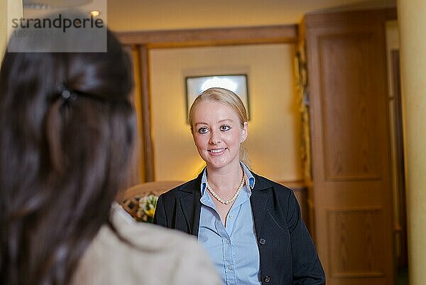 Schöne stilvolle blonde Hotelrezeptionistin steht hinter dem Serviceschalter in einer Hotellobby und schaut mit einem freundlichen Lächeln in die Kamera