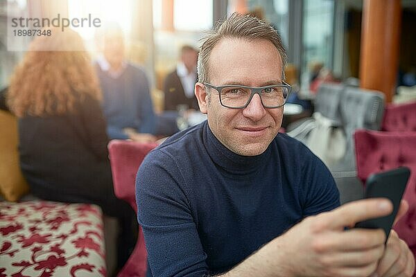 Attraktiver Mann mittleren Alters mit Brille sitzt in einem Restaurant und hält sein Handy in der Hand  während er in die Kamera lächelt