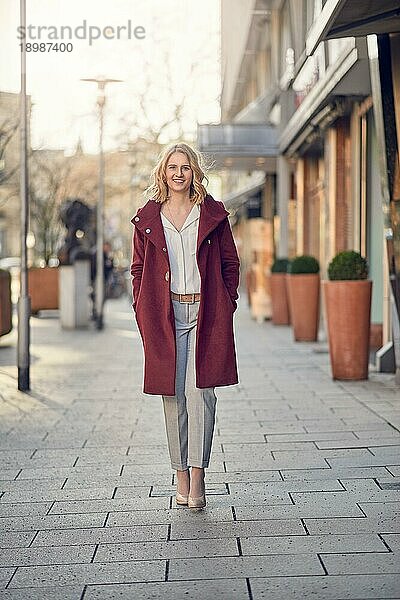 Attraktive  stilvolle junge Frau in einem kastanienbraunen Mantel  die mit den Händen in den Taschen in einer Straße der Stadt steht und in die Kamera lächelt