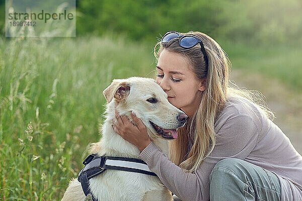 Glücklich lächelnder goldener Hund  der ein Laufgeschirr trägt und seiner hübschen jungen Besitzerin gegenübersitzt  die ihn mit einem liebevollen Lächeln im Freien auf dem Lande streichelt