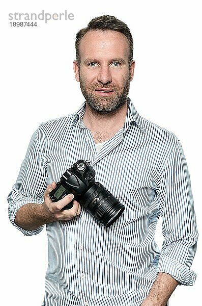 Gutaussehender Fotograf mit einem freundlichen Lächeln  der seine Kamera lachend in die Kamera hält  Oberkörperporträt in entspannter Pose auf grauem Untergrund