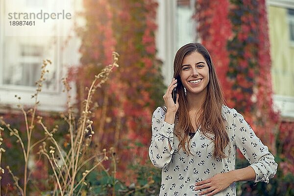Lächelnde junge Frau  die im Herbst mit einem Handy plaudert  steht vor einem weißen Haus mit einer bunten Kletterpflanze und lächelt fröhlich in die Kamera  die von der Sonne angestrahlt wird