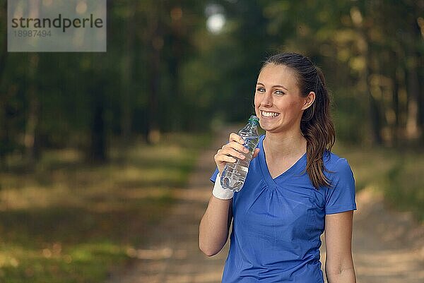 Fitte schlanke junge Frau in Sportkleidung trinkt Wasser in Flaschen  während sie auf einem Waldweg eine Pause einlegt  um während eines Trainingslaufs auf dem Lande neue Flüssigkeit zu sich zu nehmen  Gesundheits und Fitnesskonzept