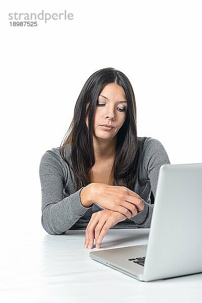 Gelangweilte attraktive junge Büroangestellte  die an ihrem Laptop sitzt und auf ihre Uhr schaut  um zu sehen  ob es Zeit ist  nach Hause zu gehen  auf weißem Grund