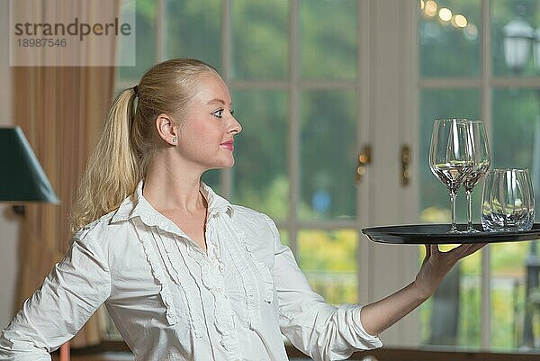 Elegante  schöne junge Frau  die Getränke serviert und dabei anmutig das Tablett mit Weißweingläsern auf ihren Fingerspitzen balanciert  im Profil stehend und mit einem freundlichen Lächeln