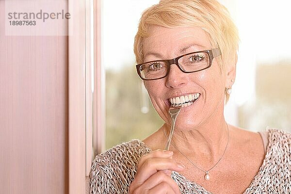 Nahaufnahme Glückliche blonde Frau mittleren Alters  die eine Brille trägt und in eine Gabel beißt  während sie in die Kamera schaut