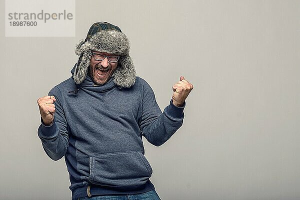 Fröhlicher Mann mit Brille und Wintermütze  der jubelnd und feiernd die geballten Fäuste in die Luft streckt  mit jubelndem Gesichtsausdruck  über grauem Hintergrund mit Kopierraum