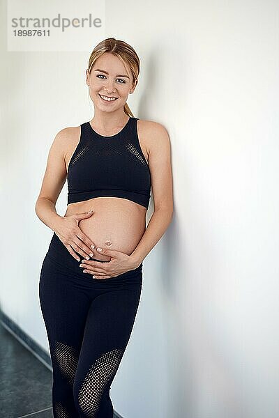 Attraktive  schwangere  freundliche junge Frau  die an eine weiße Wand gelehnt steht und ihre Hände auf ihren nackten Bauch legt und ihren Babybauch streichelt  während sie sich mit ihrem ungeborenen Kind verbindet und glücklich in die Kamera lächelt