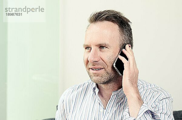 Horizontales Porträt eines gut aussehenden kaukasischen Mannes mittleren Alters  der ein langärmeliges buntes Hemd trägt  während er ein angenehmes Gespräch am Mobiltelefon führt  drinnen