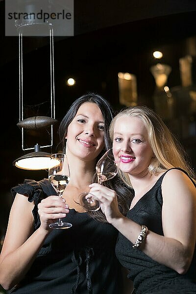 Zwei schöne  elegante Frauen in stilvollen  schlichten schwarzen Cocktailkleidern trinken ein Glas gekühlten Champagner  während sie einen gemeinsamen Abend feiern