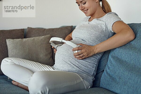 Lächelnde  glückliche  schwangere junge Frau  die ihrem ungeborenen Baby Musik mit Kopfhörern vorspielt  indem sie einen Stereokopfhörer an ihren Bauch hält  während sie im Schneidersitz auf einem Sofa sitzt