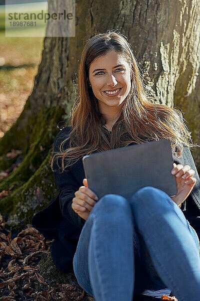 Glückliche attraktive junge Frau  die unter einem Baum sitzt  sich an den Stamm lehnt  ein Tablet auf den Knien hat und E Learning betreibt  und der Kamera ein freundliches  warmes Lächeln schenkt