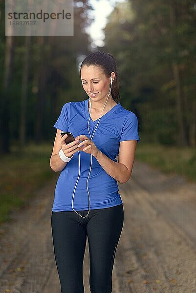 Attraktive  glückliche Frau  die mit ihrem MP3 Player und Ohrstöpseln auf einem unbefestigten Waldweg steht und mit einem Lächeln der Freude im Gesicht Musik hört