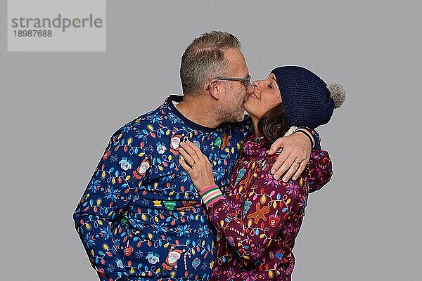 Glückliches Paar mittleren Alters  das sich küsst  während es zusammen Weihnachten feiert und in bunter  weihnachtlicher Kleidung vor einer grauen Wand posiert