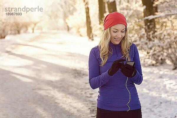 Eine Frau  die bei einem gesunden Winterspaziergang durch einen verschneiten Park in der frühen Morgensonne mit einem glücklichen Lächeln ihr Handy auf Textnachrichten überprüft