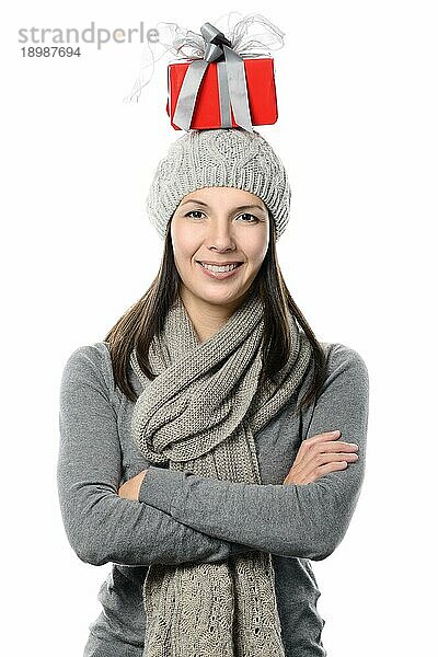 Glückliche  attraktive junge Frau in Wintermode  die ein Weihnachtsgeschenk auf dem Kopf balanciert und in die Kamera schaut  die Hände vor sich verschränkt und mit einem charmanten Lächeln  vor weißem Hintergrund