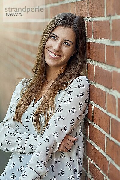 Attraktive junge Frau mit einem schönen natürlichen Lächeln  die sich an eine Außenmauer lehnt und in die Kamera blickt  mit einem Streiflicht zur Seite