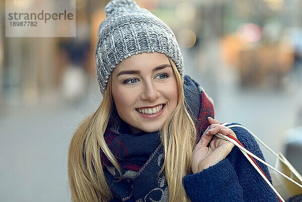 Wunderschöne junge Frau mit Weihnachtseinkäufen in einer gestrickten Wintermütze  die glücklich lächelt  während sie auf einer städtischen Straße hinter sich blickt