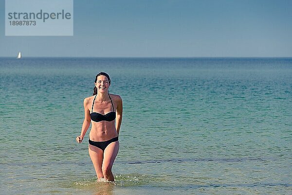 Lachende schöne junge Frau in einem schwarzen Bikini  die spazieren geht und ihr langes brünettes Haar in die Luft wirft  an einem tropischen Strand mit einem ruhigen Ozean im Hintergrund mit Kopierraum