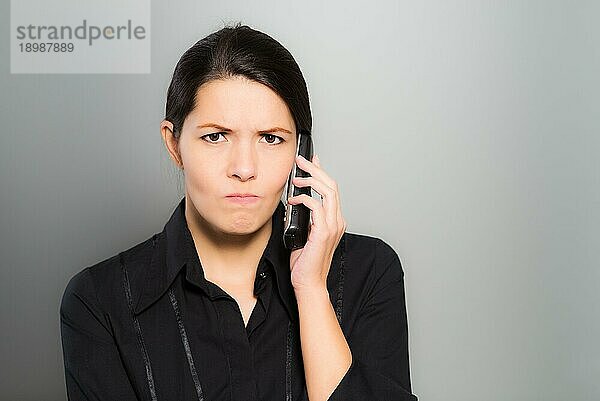 Verwirrte  schöne junge Frau  die mit ihrem Handy telefoniert und mit verwirrtem Blick und Stirnrunzeln in die Kamera schaut  vor einem grauen Studiohintergrund