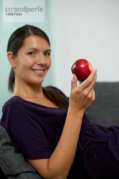 Hübsche junge Frau  die einen frischen knackigen roten Apfel in der Hand hält  während sie sich zu Hause auf dem Sofa entspannt und der Kamera ein freundliches Lächeln schenkt