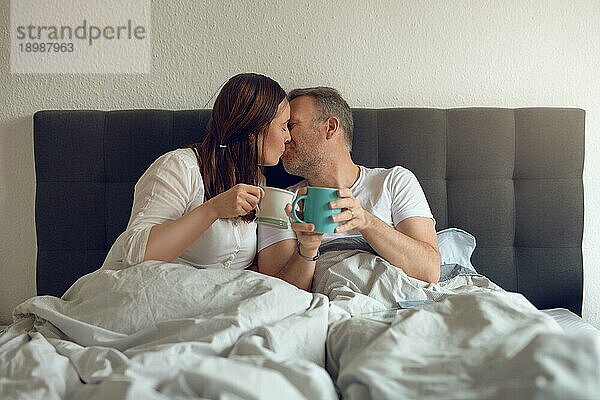 Glückliches Paar mittleren Alters  das sich morgens im Bett küsst und eine Tasse Kaffee hält