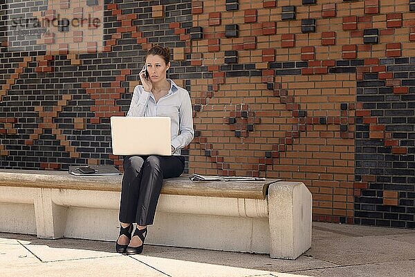 Freundliche attraktive junge Frau  die in einem Kommunikations und Mobilitätskonzept im Freien mit Kopierraum an einem Laptop arbeitet und mit einem Mobiltelefon spricht