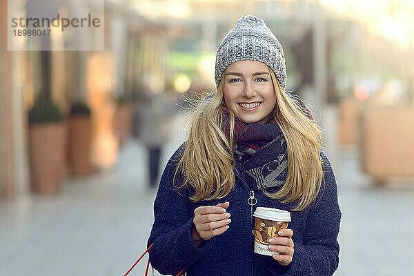 Wunderschöne junge Frau mit Weihnachtseinkäufen in einer gestrickten Wintermütze  die fröhlich lächelt  während sie auf einer städtischen Straße hinter sich blickt