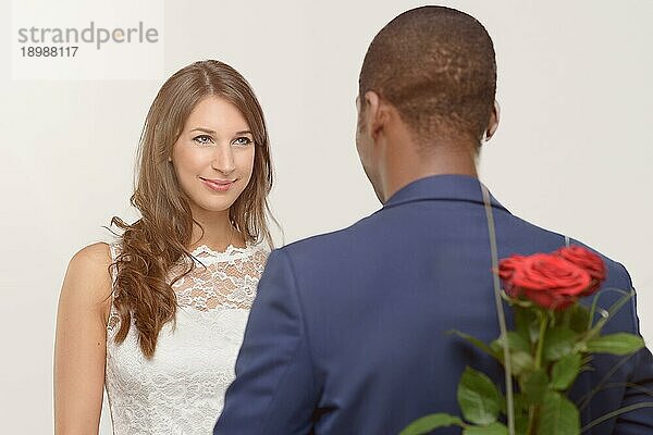Stilvoller romantischer afrikanischer Mann mit einer roten Rose  die er hinter seinem Rücken versteckt  um seine attraktive lächelnde Geliebte am Valentinstag zu überraschen