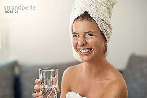 Glückliche attraktive junge Frau mit einem netten freundlichen Grinsen  die ein sauberes weißes Handtuch um ihr Haar trägt und ein Glas reines frisches Wasser hält und in die Kamera lacht