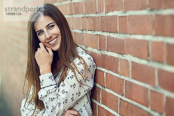 Attraktive junge Frau mit einem schönen natürlichen Lächeln  die sich an eine Außenmauer lehnt und in die Kamera blickt  mit einem Streiflicht zur Seite
